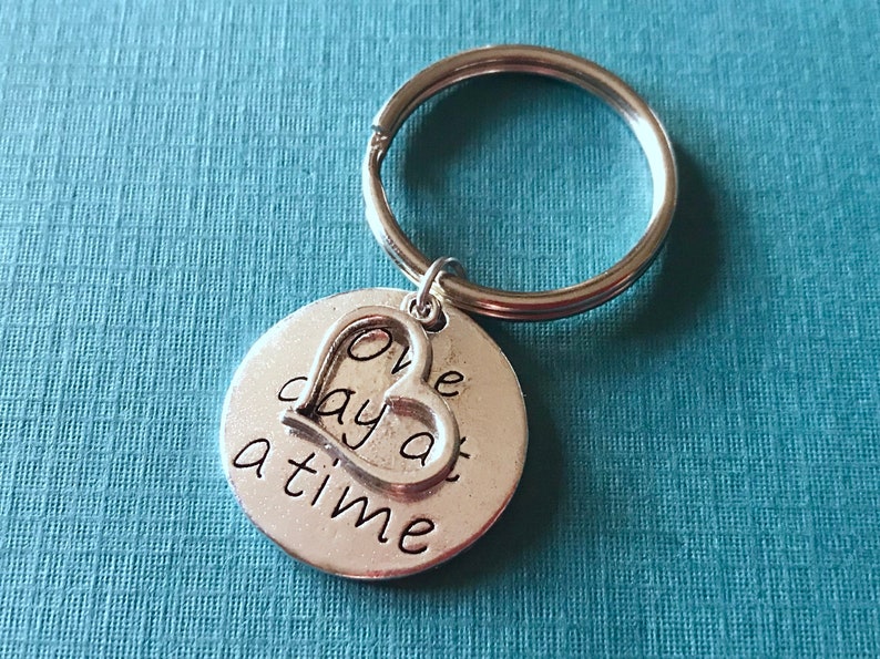 May include: A silver-colored keychain with a round charm and a heart-shaped cutout. The charm is inscribed with the words "One day at a time" in a handwritten style font. The keychain is on a blue background.