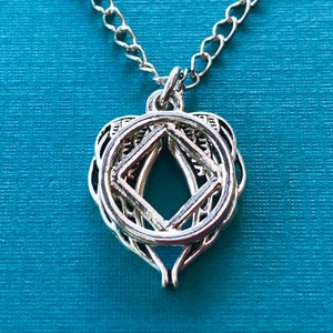 May include: Silver pendant necklace with an intricate openwork design. The pendant features a diamond shape at the centre within a circular frame, suspended from a delicate chain. The necklace is set against a teal background.