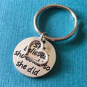 May include: Silver-colored keychain with a round charm and a heart-shaped accent. The charm has the words "She believed she could so she did" engraved on it. The keychain is on a blue background.