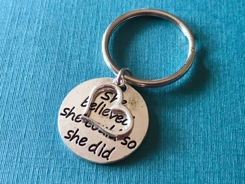 May include: Silver-colored keychain with a round charm and a heart-shaped accent. The charm has the words "She believed she could so she did" engraved on it. The keychain is on a blue background.