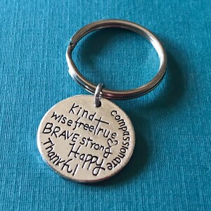 May include: A silver-colored metal keychain with a round pendant. The pendant is inscribed with words like "Kind," "Brave," and "Happy." The keychain ring is silver and attached to the pendant. The background is a solid teal color.