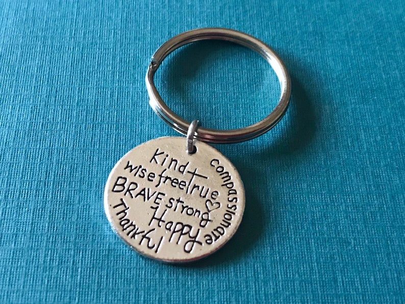 May include: A silver-colored metal keychain with a round pendant. The pendant is inscribed with words like "Kind," "Brave," and "Happy." The keychain ring is silver and attached to the pendant. The background is a solid teal color.