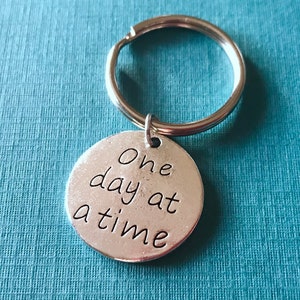 May include: A silver-colored keychain with a round pendant. The pendant has the phrase "One day at a time" in a handwritten style font. The keychain ring is silver and attached to the pendant.