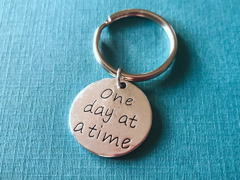 May include: A silver-colored keychain with a round pendant. The pendant has the phrase "One day at a time" in a handwritten style font. The keychain ring is silver and attached to the pendant.