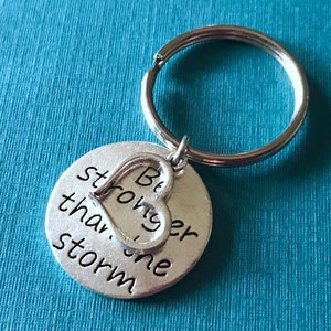 May include: A silver-colored keychain with a round pendant and a heart-shaped charm. The pendant has the words "Be stronger than the storm" in black lettering. The keychain is attached to a silver key ring.