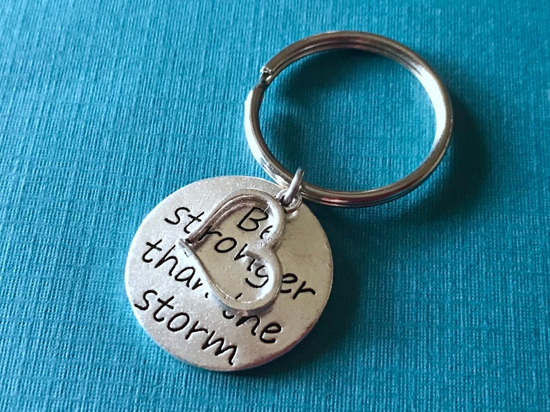 May include: A silver-colored keychain with a round pendant and a heart-shaped charm. The pendant has the words "Be stronger than the storm" in black lettering. The keychain is attached to a silver key ring.
