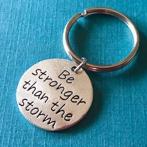 May include: A silver-colored keychain with a round pendant. The pendant has the phrase "Be stronger than the storm" in black lettering. The keychain ring is silver and attached to the pendant.