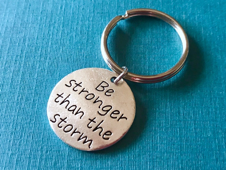 May include: A silver-colored keychain with a round pendant. The pendant has the phrase "Be stronger than the storm" in black lettering. The keychain ring is silver and attached to the pendant.