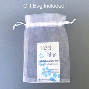 May include: A sheer white organza gift bag with a drawstring closure. A rectangular label with the words "hazel blue" and "inspirational jewelry" is visible. The bag is on a gray background. Text at the top reads "Gift Bag Included!"