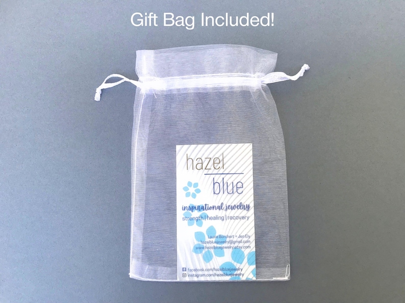 May include: A sheer white organza gift bag with a drawstring closure. A rectangular label with the words "hazel blue" and "inspirational jewelry" is visible. The bag is on a gray background. Text at the top reads "Gift Bag Included!"