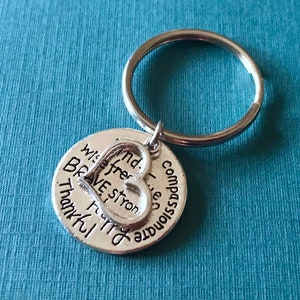 May include: Silver-colored keychain with a round charm and a heart-shaped cutout. The charm is inscribed with words like "brave," "strong," and "thankful." The keychain is on a blue background.