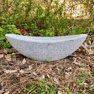 Op de afbeelding: Een grijze betonnen plantenbak in de vorm van een boot. De plantenbak heeft een ruw, getextureerd oppervlak en staat op een bed van dennennaalden en bladeren.