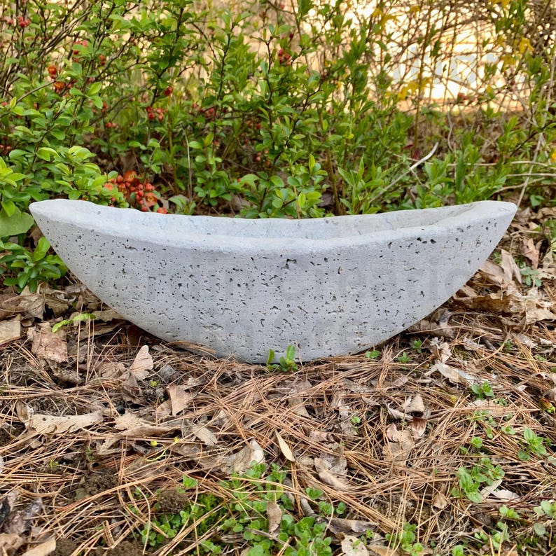 Op de afbeelding: Een grijze betonnen plantenbak in de vorm van een boot. De plantenbak heeft een ruw, getextureerd oppervlak en staat op een bed van dennennaalden en bladeren.