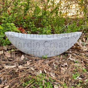 Op de afbeelding: Een grijze betonnen trogplanter met een ruw, getextureerd oppervlak. De planter is bootvormig en staat op een bed van dennennaalden.
