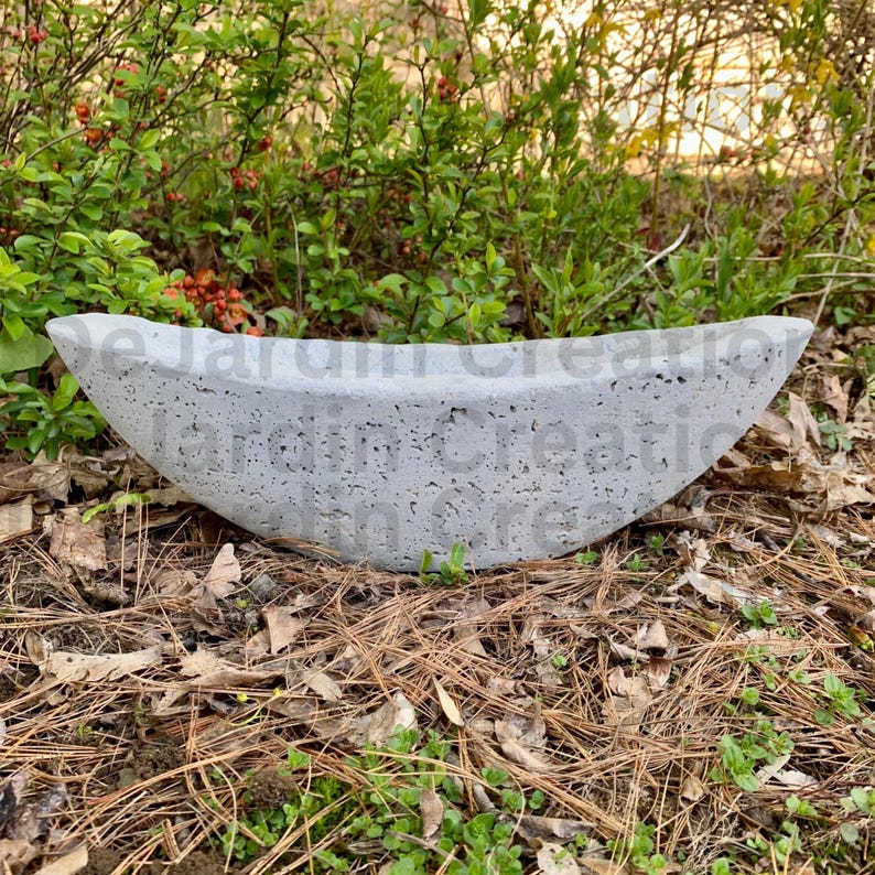 Op de afbeelding: Een grijze betonnen trogplanter met een ruw, getextureerd oppervlak. De planter is bootvormig en staat op een bed van dennennaalden.