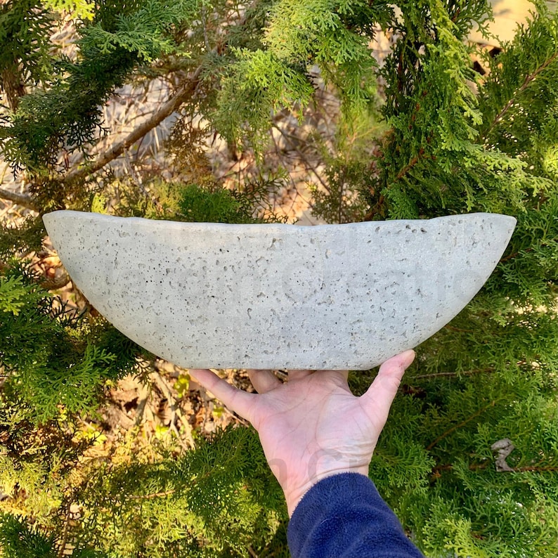Op de afbeelding: Een grote, ovale betonnen plantenbak met een ruw, getextureerd oppervlak. De plantenbak is lichtgrijs van kleur en heeft een licht gebogen vorm.