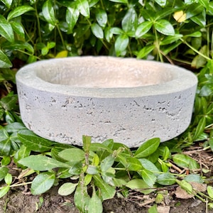 May include: A circular, light gray concrete bowl with a porous, textured exterior surface and a smooth, wide rim. The vessel features a shallow, open interior basin and a flat base, showcasing a raw, industrial aesthetic with visible air pockets.