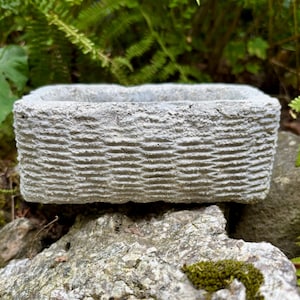 May include: A rectangular concrete planter with a rough, textured exterior surface featuring a repeating horizontal pattern of small, raised ridges. The planter has a thick, solid rim and a deep, open interior cavity for holding plants or soil.