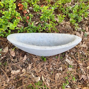 Op de afbeelding: Een grijze betonnen plantenbak in de vorm van een boot. De plantenbak staat op een bed van dennennaalden en bruine bladeren.