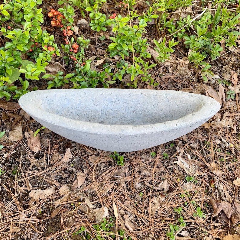 Op de afbeelding: Een grijze betonnen plantenbak in de vorm van een boot. De plantenbak staat op een bed van dennennaalden en bruine bladeren.