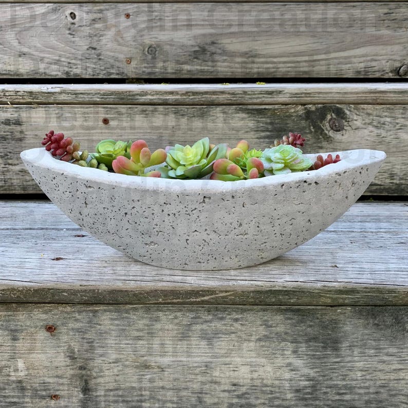 Op de afbeelding: Een grijze betonnen plantenbak in de vorm van een boot, gevuld met groene en rode succulenten.