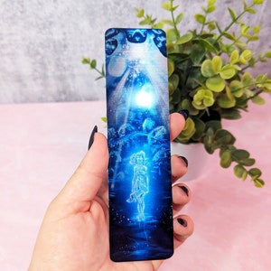 May include: A blue and silver bookmark with a woman standing in the center. The woman is outlined in white and is surrounded by a blue and silver background. The bookmark is made of metal and has a shiny finish.