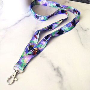 K&ouml;nnte beinhalten: Ein lilafarbenes Lanyard mit einem bunten Spr&uuml;hfarben-Design und Comicfiguren. Das Lanyard hat einen silbernen Metallclip.