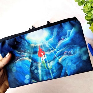 K&ouml;nnte beinhalten: Eine schwarze Canvas-Rei&szlig;verschlusstasche mit einer farbenfrohen Illustration von Arielle, der kleinen Meerjungfrau, die unter Wasser schwimmt. Die Illustration zeigt eine blaue, gr&uuml;ne und violette Ozeanszene mit Blasen und Licht, das von oben nach unten scheint.