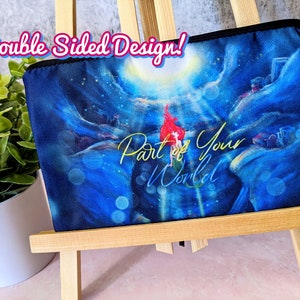 K&ouml;nnte beinhalten: Eine blaue und schwarze Rei&szlig;verschlusstasche mit einem farbenfrohen Bild einer Meerjungfrau und dem Text "Part of Your World". Die Tasche wird auf einem Holzst&auml;nder pr&auml;sentiert. Doppelseitiges Design!