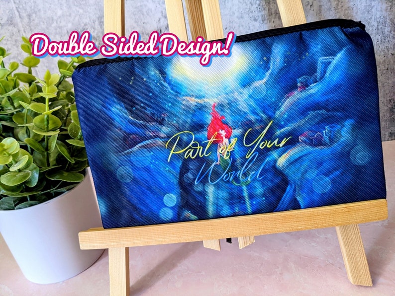 K&ouml;nnte beinhalten: Eine blaue und schwarze Rei&szlig;verschlusstasche mit einem farbenfrohen Bild einer Meerjungfrau und dem Text "Part of Your World". Die Tasche wird auf einem Holzst&auml;nder pr&auml;sentiert. Doppelseitiges Design!