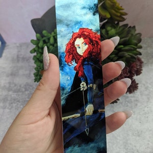 May include: A rectangular bookmark featuring a watercolor illustration of a character with fiery red hair, blue eyes, and a bow and arrow. The background has a blue and white cloudy sky.