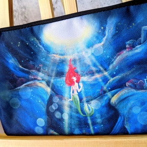 K&ouml;nnte beinhalten: Eine blaue und schwarze Stofftasche mit einem aufgedruckten Bild einer rothaarigen Meerjungfrau mit einem gr&uuml;nen Schwanz, die nach oben zu einem hellen Licht schwimmt. Der Hintergrund ist dunkelblau mit einem Sternenhimmel und einem Mond.