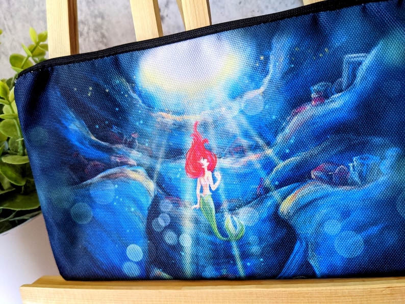 K&ouml;nnte beinhalten: Eine blaue und schwarze Stofftasche mit einem aufgedruckten Bild einer rothaarigen Meerjungfrau mit einem gr&uuml;nen Schwanz, die nach oben zu einem hellen Licht schwimmt. Der Hintergrund ist dunkelblau mit einem Sternenhimmel und einem Mond.