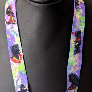 K&ouml;nnte beinhalten: Ein lilafarbenes Lanyard mit einem bunten Spritzmuster. Das Lanyard zeigt verschiedene Disney-Schurken, darunter Ursula, Dschafar und Dr. Facilier.