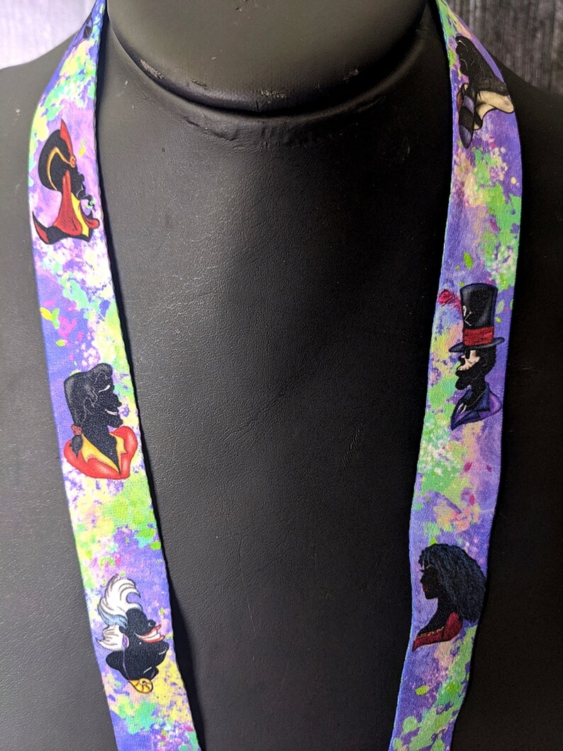 K&ouml;nnte beinhalten: Ein lilafarbenes Lanyard mit einem bunten Spritzmuster. Das Lanyard zeigt verschiedene Disney-Schurken, darunter Ursula, Dschafar und Dr. Facilier.