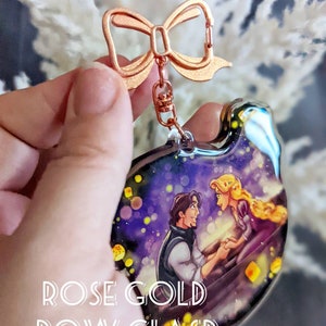 May include: A clear acrylic keychain featuring a romantic scene with a man and woman in a boat under a starry sky. The keychain has a rose gold bow clasp and the text "ROSE GOLD BOW CLASP".