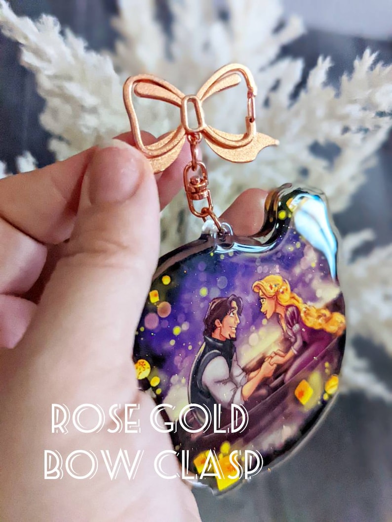 May include: A clear acrylic keychain featuring a romantic scene with a man and woman in a boat under a starry sky. The keychain has a rose gold bow clasp and the text "ROSE GOLD BOW CLASP".