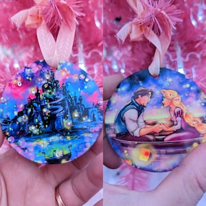 May include: Two round Christmas ornaments with a Disney theme. The first ornament features a blue and purple castle with sparkling lights. The second ornament features a scene from the movie Tangled, with Rapunzel and Flynn Rider in a boat.