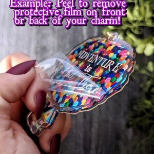 Puede incluir: Un amuleto de acr&iacute;lico transparente con forma de globo aerost&aacute;tico con las palabras "La aventura est&aacute; ah&iacute; fuera" y globos de colores. El amuleto est&aacute; siendo sostenido por una mano y se est&aacute; despegando la pel&iacute;cula protectora.