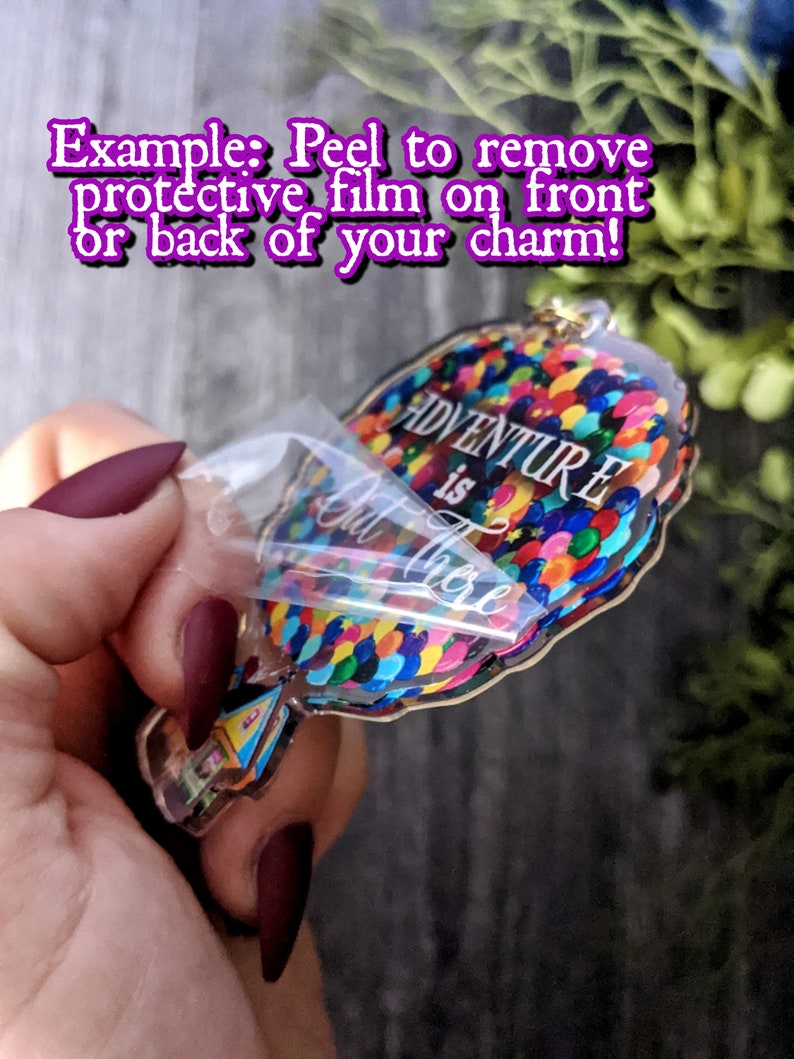 Puede incluir: Un amuleto de acr&iacute;lico transparente con forma de globo aerost&aacute;tico con las palabras "La aventura est&aacute; ah&iacute; fuera" y globos de colores. El amuleto est&aacute; siendo sostenido por una mano y se est&aacute; despegando la pel&iacute;cula protectora.