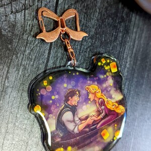 May include: A clear acrylic keychain featuring a romantic scene with a man and woman in a boat, surrounded by floating lanterns. The keychain has a copper-colored bow-shaped clasp and a small chain.
