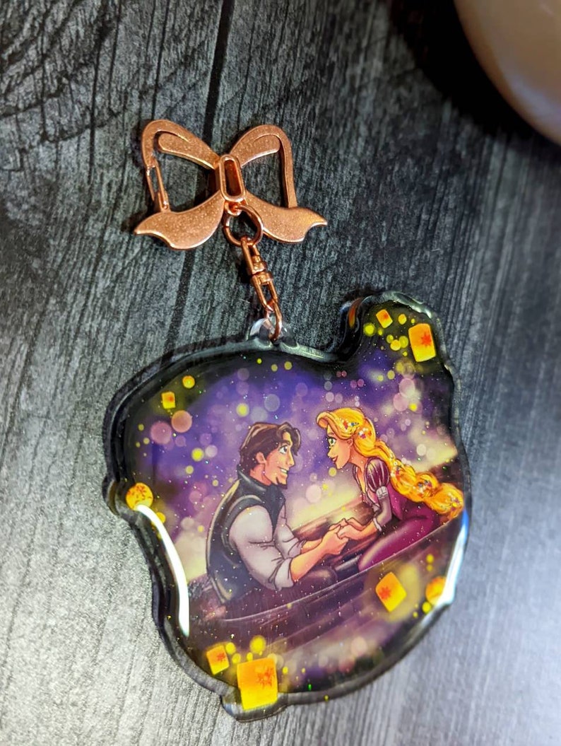 May include: A clear acrylic keychain featuring a romantic scene with a man and woman in a boat, surrounded by floating lanterns. The keychain has a copper-colored bow-shaped clasp and a small chain.