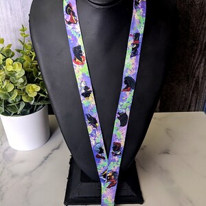 K&ouml;nnte beinhalten: Ein lilafarbenes Lanyard mit einem bunten Spritzmuster, das verschiedene Comicfiguren zeigt. Das Lanyard hat einen silbernen Metallclip.