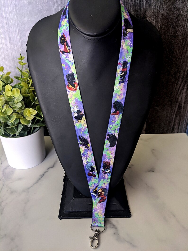 K&ouml;nnte beinhalten: Ein lilafarbenes Lanyard mit einem bunten Spritzmuster, das verschiedene Comicfiguren zeigt. Das Lanyard hat einen silbernen Metallclip.