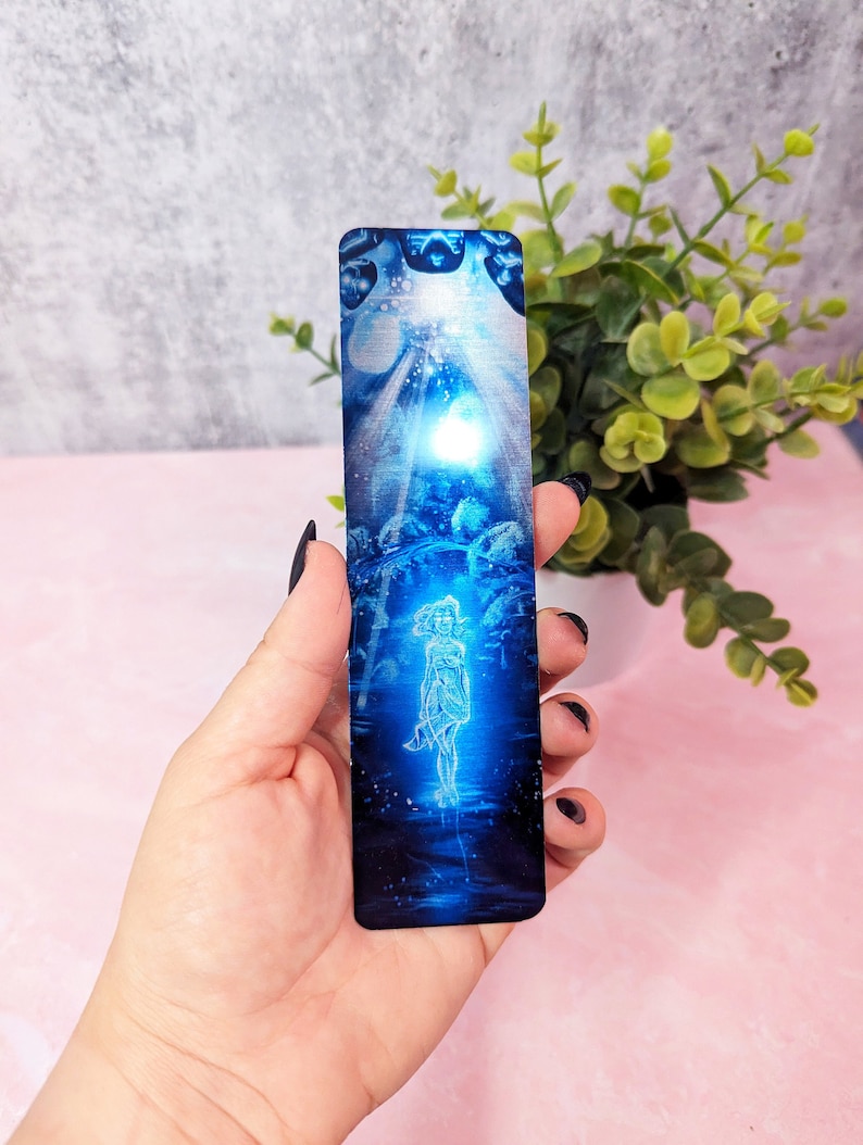 May include: A blue and silver bookmark with a woman standing in the center. The woman is outlined in white and is surrounded by a blue and silver background. The bookmark is made of metal and has a shiny finish.