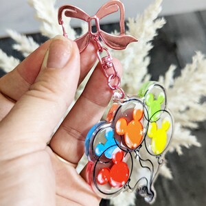 Puede incluir: Un llavero con cierre de lazo rosa y un dije transparente con cinco globos de Mickey Mouse de colores. Los globos son rojo, naranja, amarillo, verde y azul.