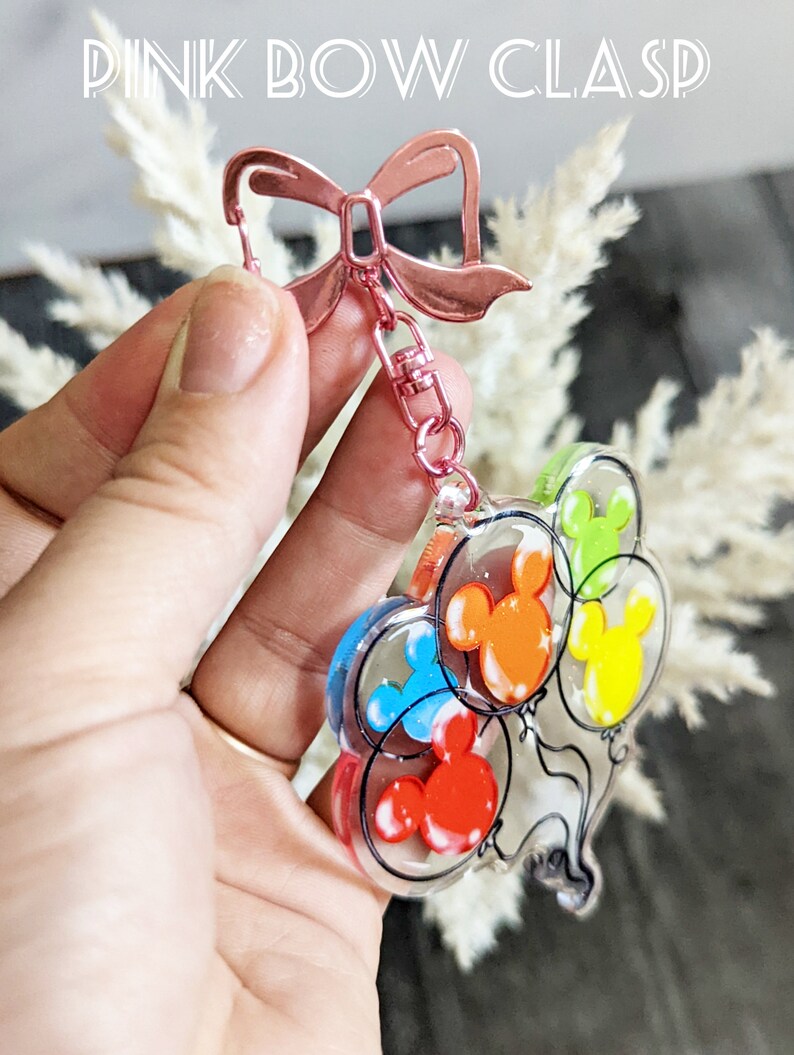 Puede incluir: Un llavero con cierre de lazo rosa y un dije transparente con cinco globos de Mickey Mouse de colores. Los globos son rojo, naranja, amarillo, verde y azul.