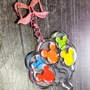 Puede incluir: Un llavero de acr&iacute;lico transparente con un lazo rosa y una cadena. El llavero presenta un grupo de cuatro globos con forma de cabeza de Mickey Mouse en diferentes colores: rojo, naranja, amarillo y azul.