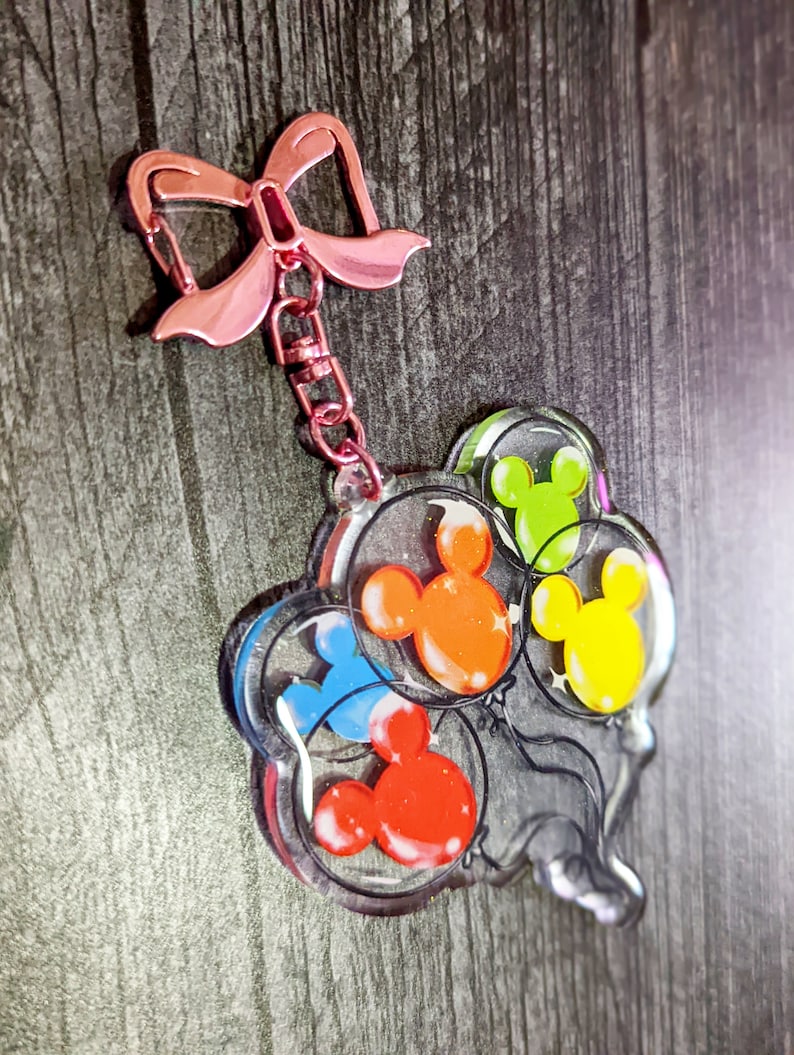 Puede incluir: Un llavero de acr&iacute;lico transparente con un lazo rosa y una cadena. El llavero presenta un grupo de cuatro globos con forma de cabeza de Mickey Mouse en diferentes colores: rojo, naranja, amarillo y azul.
