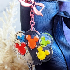 Puede incluir: Un llavero de acr&iacute;lico transparente con un lazo rosa y una cadena. El llavero presenta un grupo de globos de Mickey Mouse en rojo, naranja, amarillo, verde y azul.